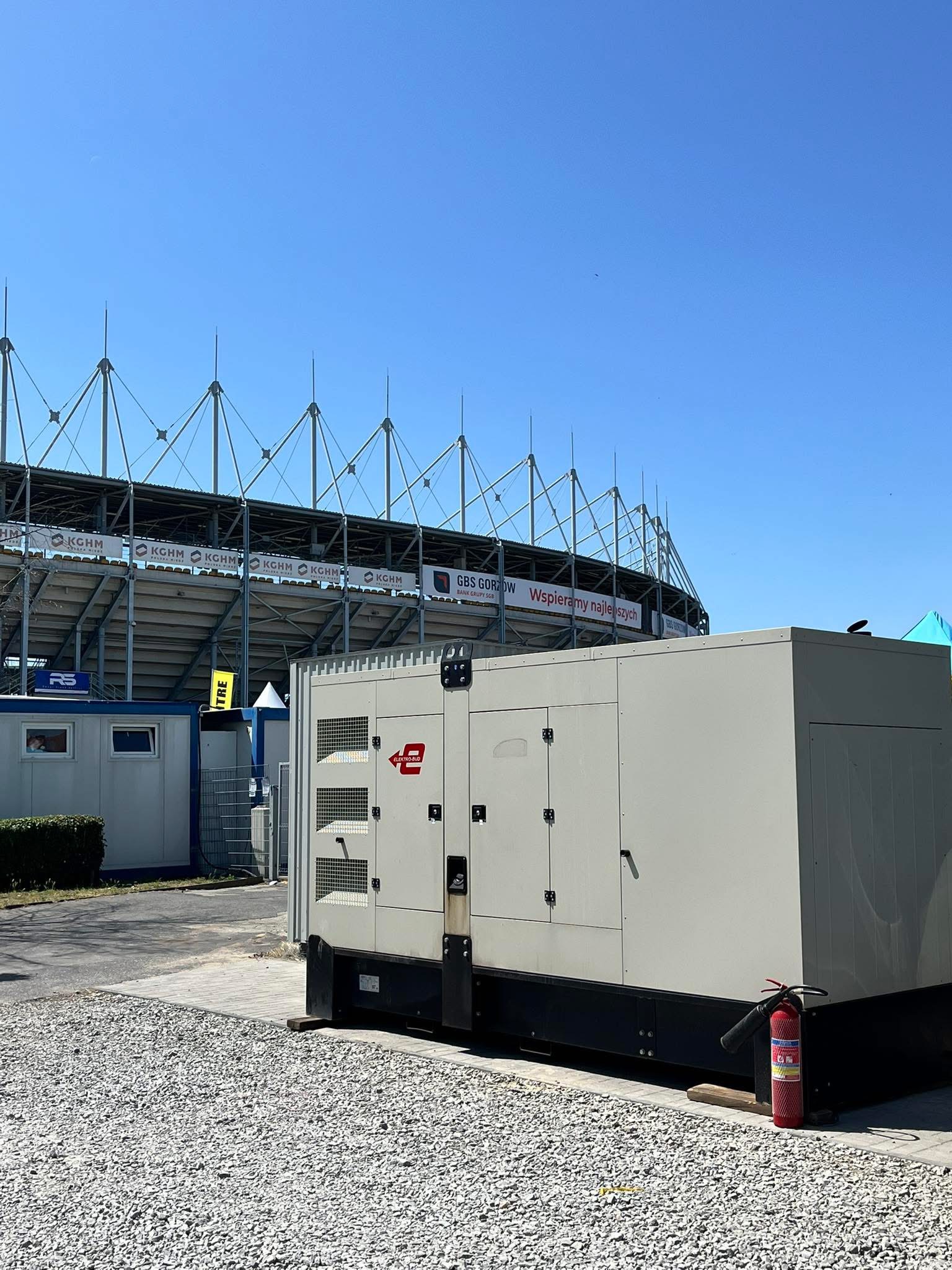 Agregat prądotwórczy Elektro-Bud ustawiony na utwardzonym terenie w pobliżu stadionu i kontenerów technicznych